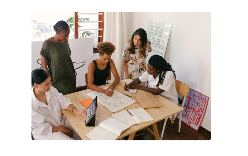 Diverso grupo de mujeres emprendedoras discutiendo perspectivas del mercado objetivo para el crecimiento del comercio electrónico alrededor de una mesa de madera.